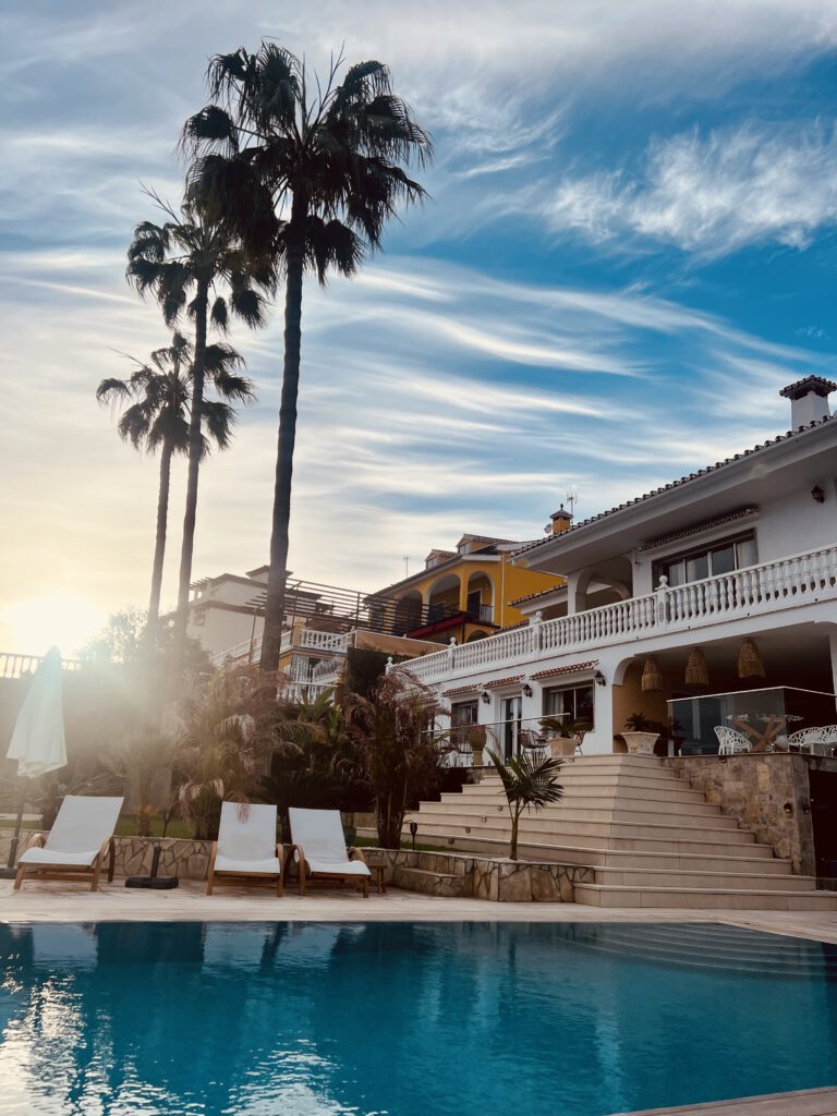 Yoga Retreat Spanien. Großer, blauer Swimmingpool der Villa Soluna in Andalusien mit weißen Sonnenliegen vor der hellen Fassade des Hauses bei strahlendem Sonnenschein.