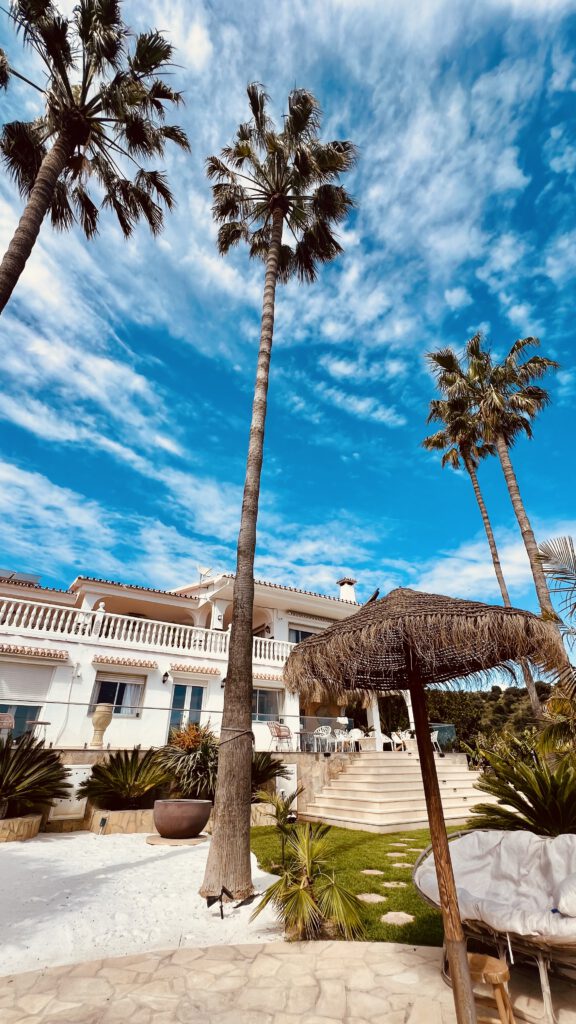 Yoga Retreat Spanien. Mediterraner Garten der Villa Soluna in Andalusien mit großen Palmen, weißem Sand und einem Stroh-Sonnenschirm unter blauem Himmel.