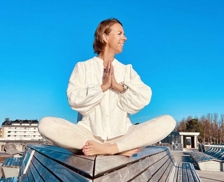 Eine lächelnde Frau (Tina von Yogacoast) sitzt im Schneidersitz in einer Yoga-Pose auf einem Holzdeck an der Ostsee in Haffkrug.