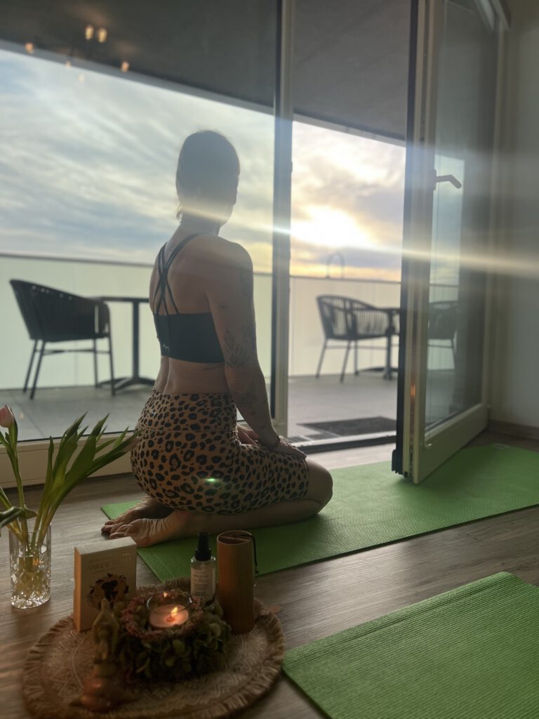 Eine Frau kniet in einer Yoga-Haltung rücklings auf einer grünen Matte in einem hellen Zimmer vor einer geöffneten Balkontür mit Blick auf die Ostsee im sanften Gegenlicht der Sonne.