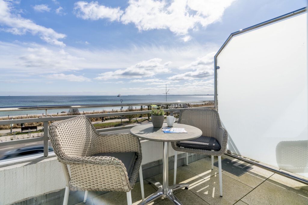 Ein Balkon mit zwei hellen Flechtsesseln und einem runden Tisch mit Blick über die Strandpromenade direkt auf die blaue Ostsee unter leicht bewölktem Himmel.