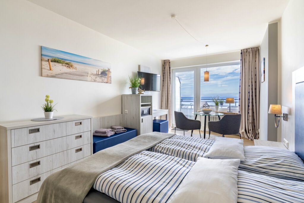 Ein helles, modern eingerichtetes Schlafzimmer mit einem großen Doppelbett in blau-weiß gestreifter Bettwäsche, einem Fernseher an der Wand und einer Fensterfront, die auf einen Balkon mit Blick zum Meer führt.