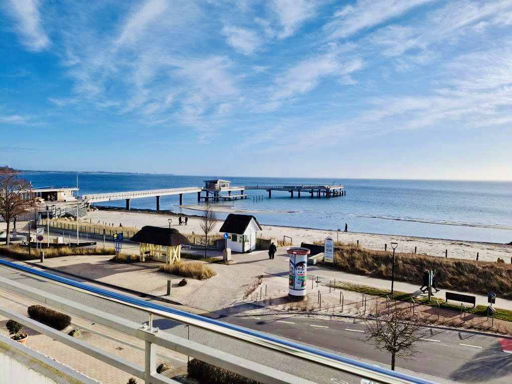 Blick von oben auf die moderne Seebrücke in Haffkrug, die weit in die blaue Ostsee hineinragt, mit der davorliegenden Strandpromenade und dem Sandstrand.