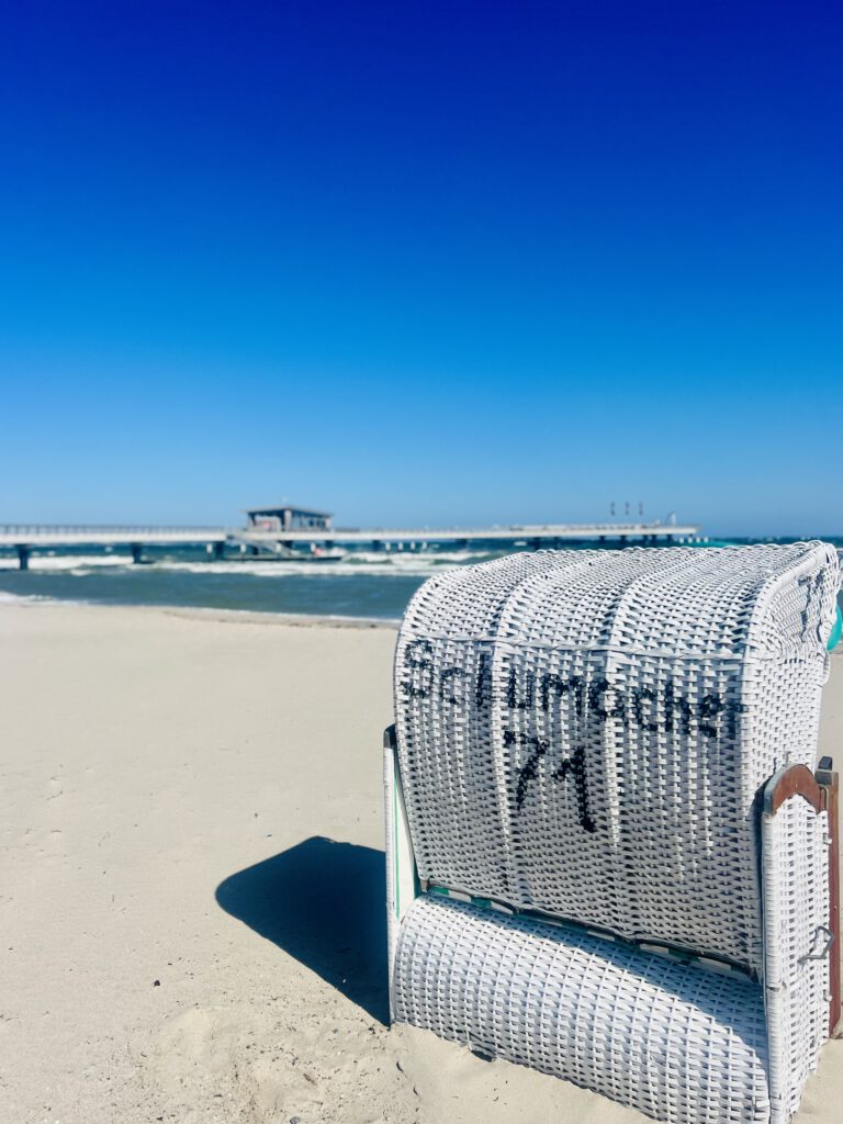 Ein weißer Strandkorb mit der Aufschrift „Schumacher 71“ steht im feinen Sandstrand von Haffkrug vor blauem Himmel, im Hintergrund ist die Seebrücke im Meer zu sehen.