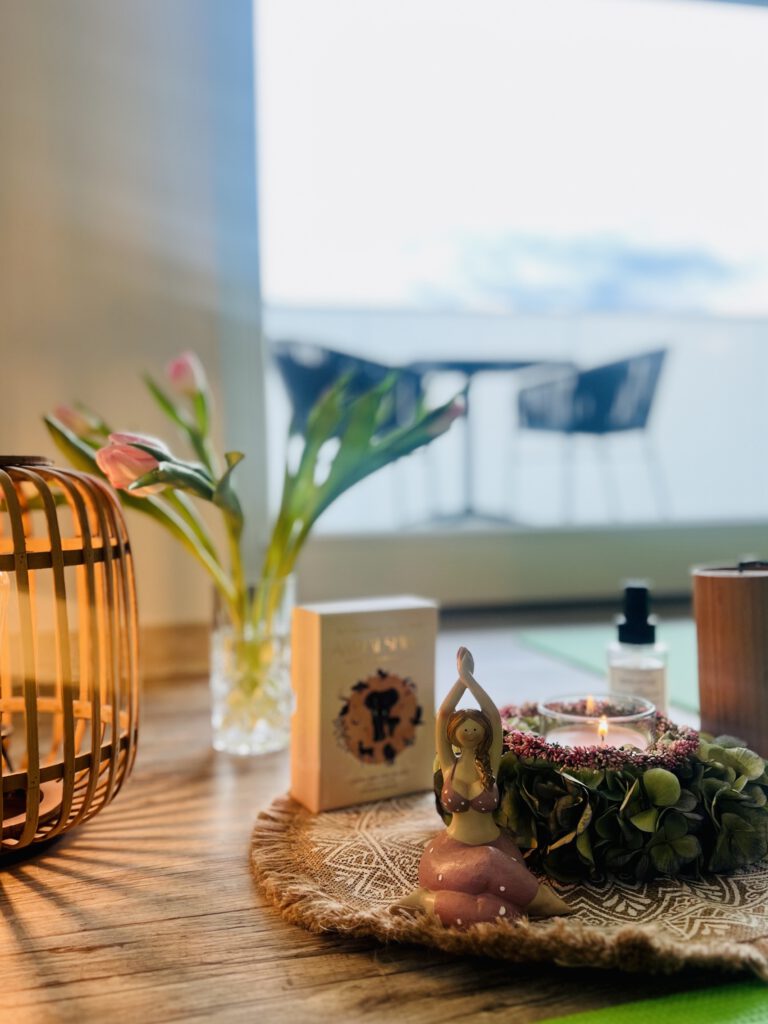 Nahaufnahme eines hölzernen Bodens mit einer kleinen Yoga-Figur, einer brennenden Kerze in einem Kranz, einer Vase mit rosa Tulpen und einer dekorativen Holzlaterne vor einer hellen Fensterfront.
