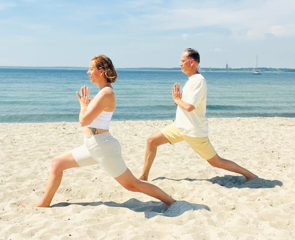 Yoga Retreat an der Ostsee. Eine Frau und ein Mann in der Krieger-Pose mit den Händen vor der Brust (Anjali Mudra) am Strand beim Yoga Retreat an der Ostsee.