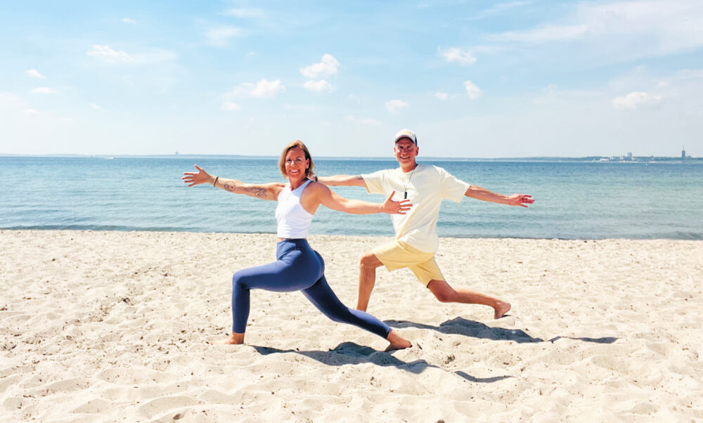 Dynamische Yogaübung im Sand von Scharbeutz. Der Blick geht direkt nach vorne, im Hintergrund erstreckt sich die ruhige Ostsee unter blauem Himmel.