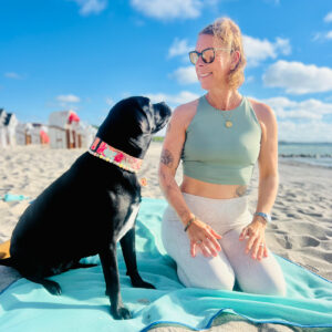 Yoga am Strand von Scharbeutz mit Hund: Eine Frau von Yogacoast praktiziert Strandyoga mit ihrem schwarzen Hund (Yogadog) an der Ostsee.