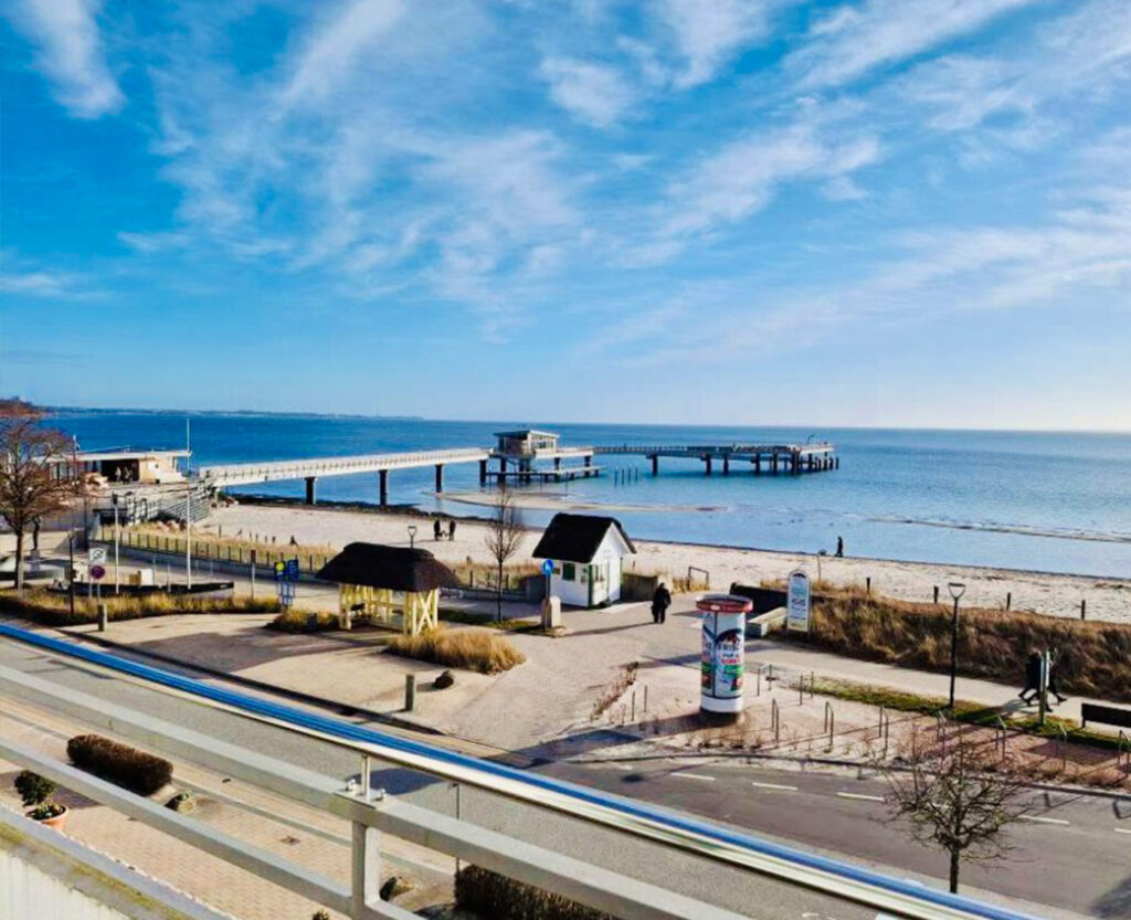 Aussicht von Balkon des Apartments strandallee.net auf die Seebrücke, den Ostseestrand und das Meer in Scharbeutz Haffkrug bei blauem Himmel.