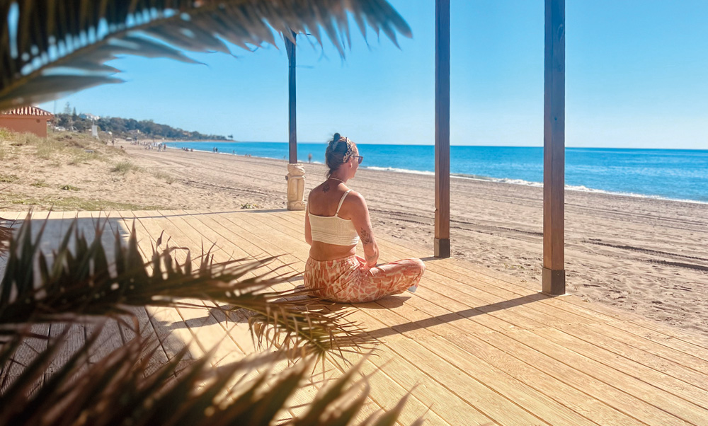 Tina von YOGACOAST sitzt auf einer Holzterrasse am Strand im Meditationssitz mit Blick auf das Meer bei einem Yoga Retreat in Andalusien.