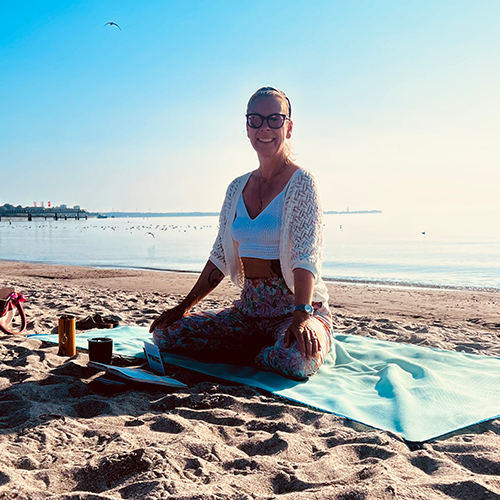 Tina von YOGACOAST sitzt im Lotussitz auf einer hellblauen Yogamatte am Sandstrand vor ruhiger Seekulisse bei Sonnenschein.