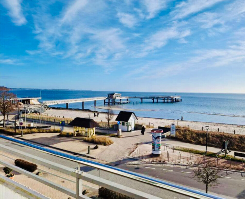 Aussicht von Balkon des Apartments strandallee.net auf die Seebrücke, den Ostseestrand und das Meer in Scharbeutz Haffkrug bei blauem Himmel.
